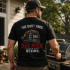 Firefighter dad wearing a black t-shirt with back print featuring a firefighter helmet and cap graphic with the text "The Shift Ends. Dad Mode Begins." while grilling outdoors in a backyard family setting.